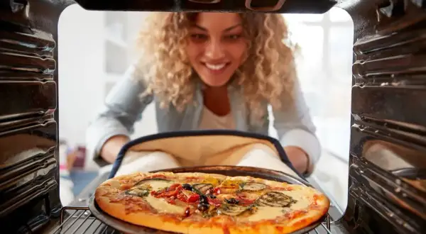 Uśmiechnięta kobieta wyjmuje z piekarnika gorącą pizzę. Idealna temperatura pieczenia pizzy to klucz do sukcesu!