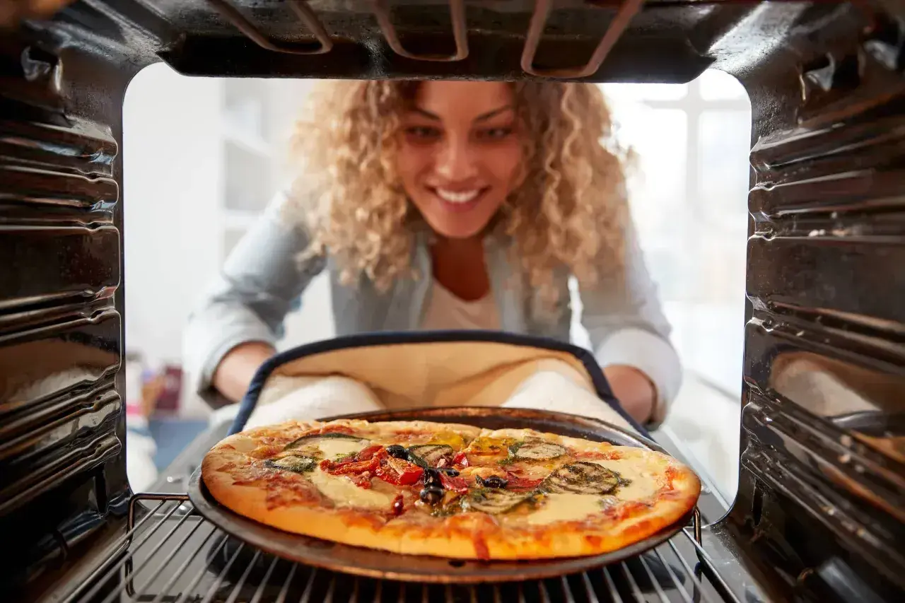 Uśmiechnięta kobieta wyjmuje z piekarnika gorącą pizzę. Idealna temperatura pieczenia pizzy to klucz do sukcesu!