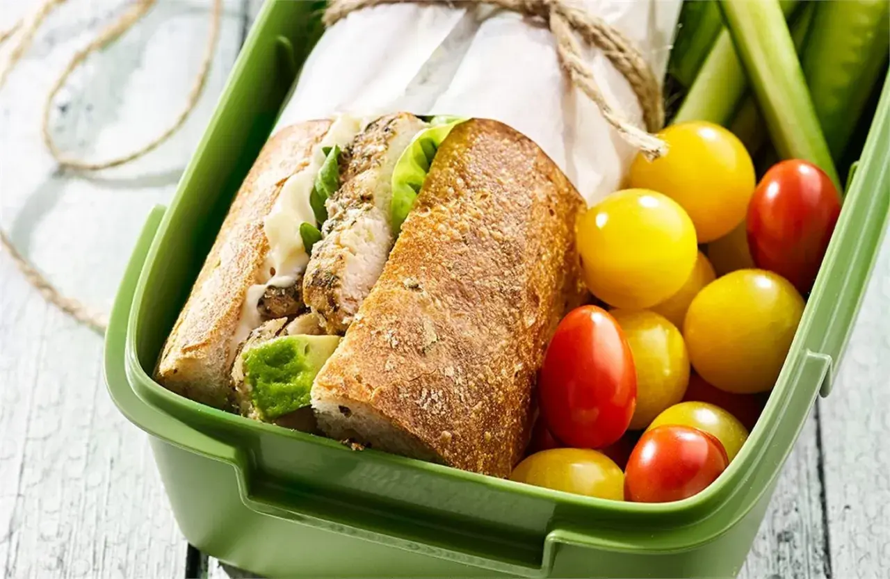 Pyszna kanapka z kurczakiem w zielonym pudełku, obok pomidorki i ogórki. Idealna na lunch!