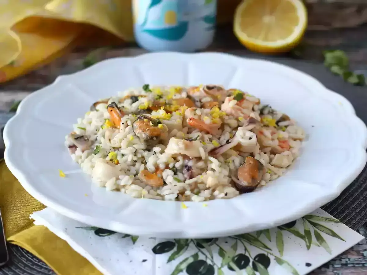 Pyszne risotto z owocami morza, udekorowane skórką cytrynową i ziołami, na białym talerzu.