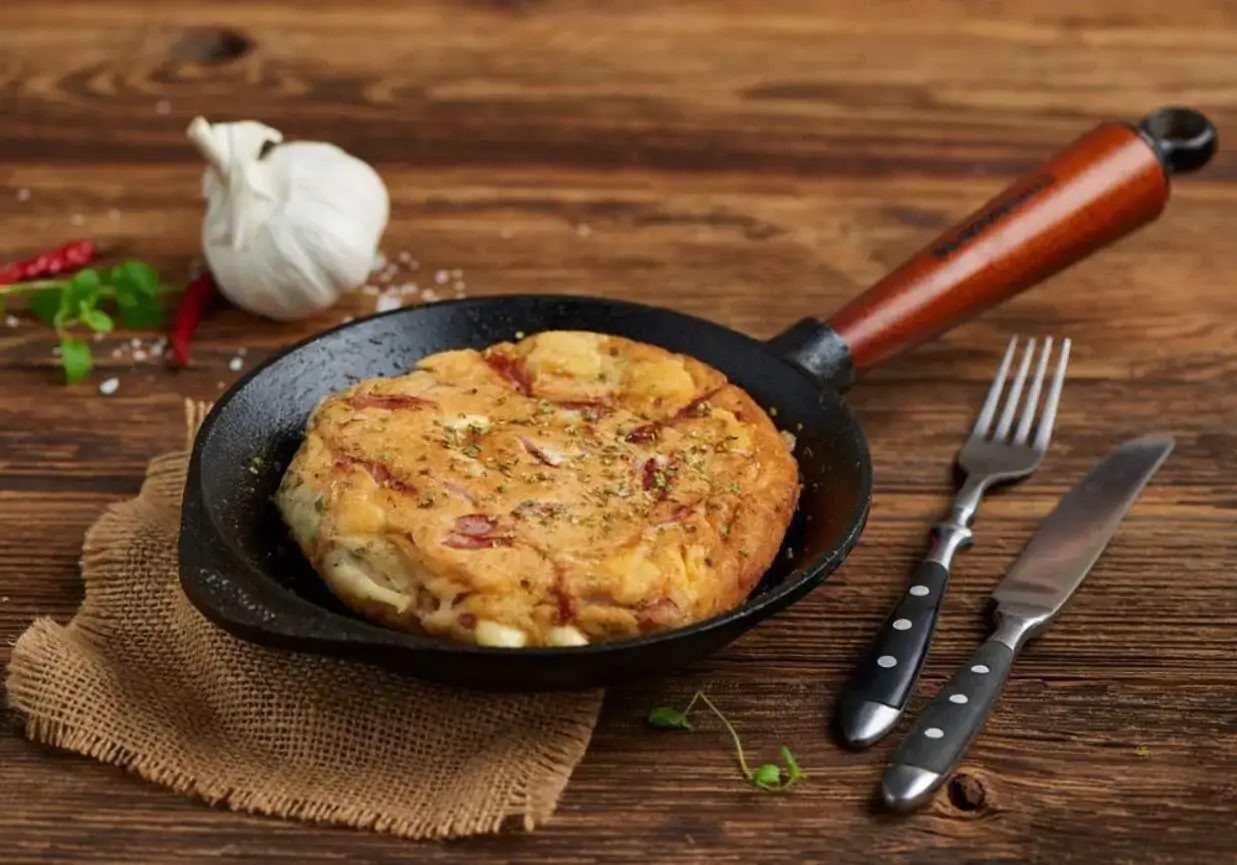 Pyszny omlet z szynką i serem, podany na patelni, z widelcem i nożem obok.