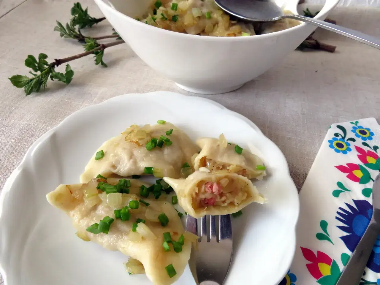 Pyszne pierogi z ziemniakami i boczkiem, posypane cebulką i szczypiorkiem, na białym talerzu.