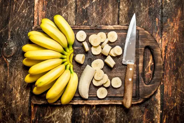 Pęczek bananów, pokrojone plasterki i nóż na drewnianej desce. Czy banany można trzymać w lodówce?