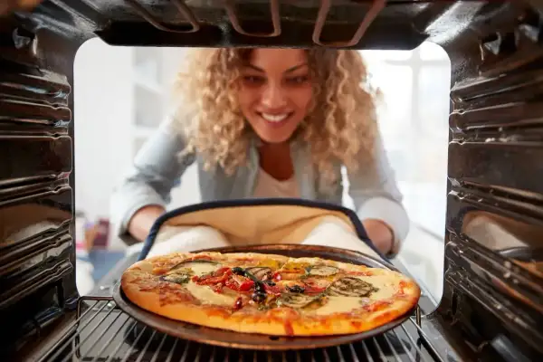 Uśmiechnięta kobieta wyjmuje z piekarnika gorącą pizzę. Idealna temperatura pieczenia pizzy to klucz do sukcesu!